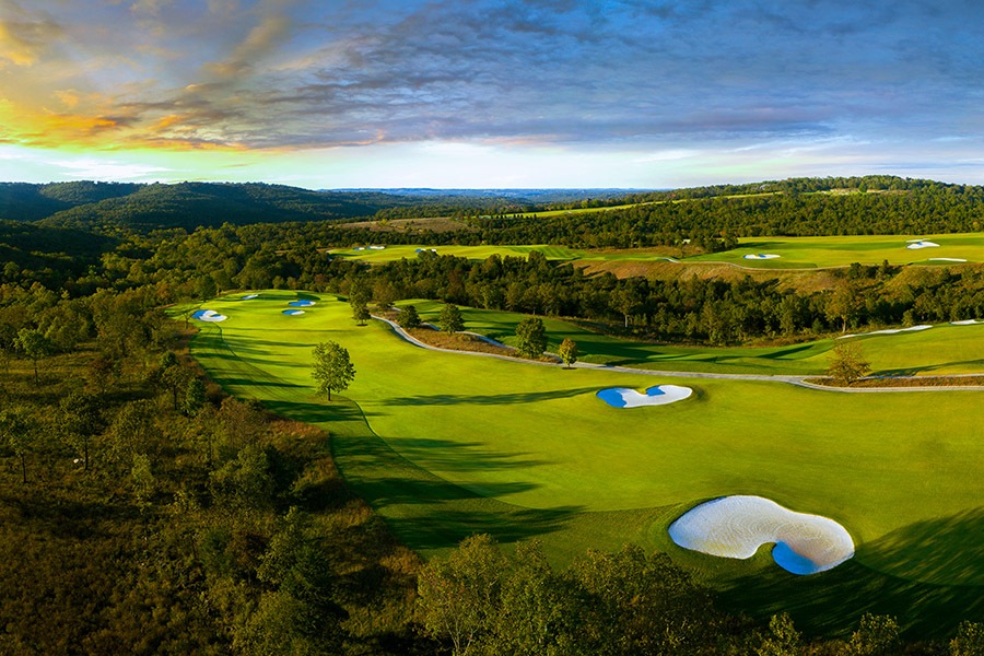 a fairway with vibrant green grass splits between dense forest. deep shadows lay across the fairway from trees