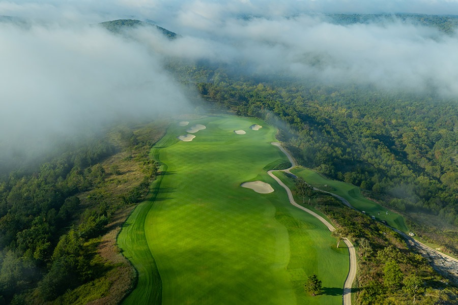 a straight fairway from a birds eye view shows a checked mowing pattern on the freshly cut grass. low fog surrounds the hole