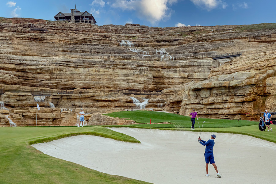 tigers woods swinging in a sand bunker while other golfers watch at the bottom of large rock formation