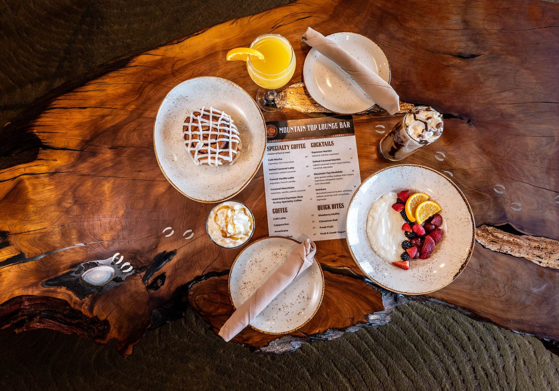 several breakfast items including a cinnamon roll with a beautiful criss cross icing pattern is joined by orange juice, fruits and yogurt on a wooden table