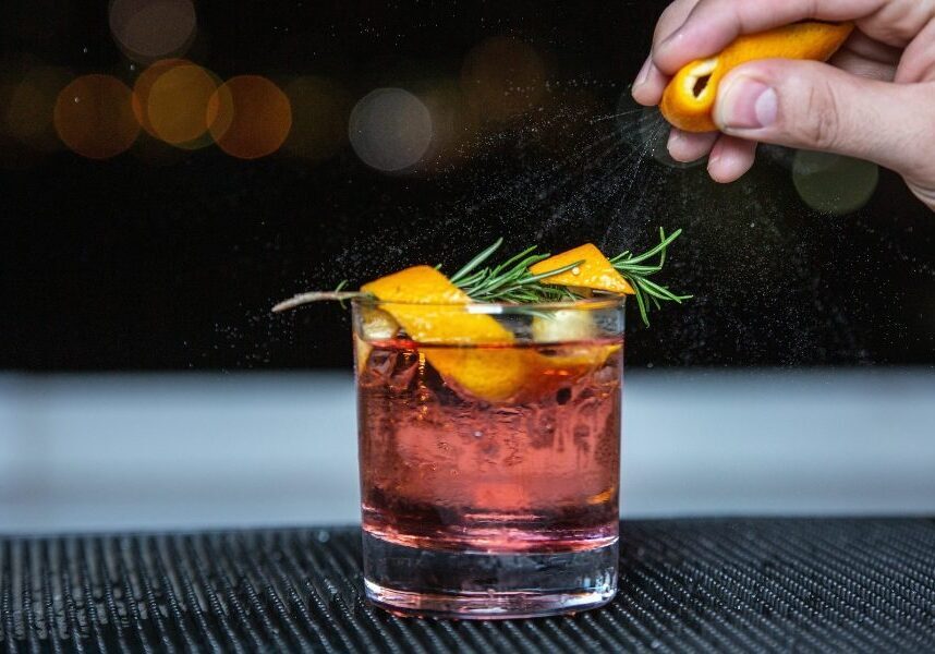 A bartender garnishes a cocktail with orange mist and rosemary.