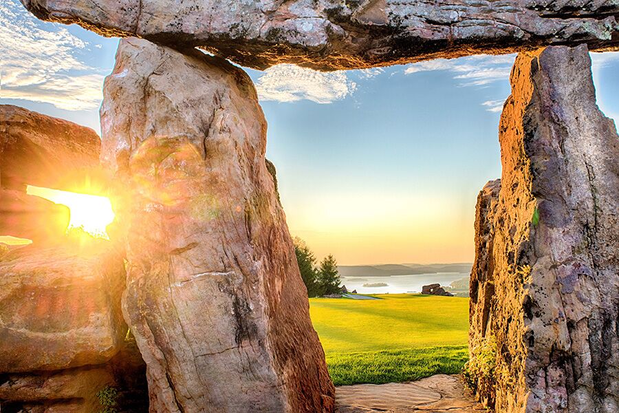 three large rocks create an archway that lead to a golf course where a sunset covers the hills with a golden light