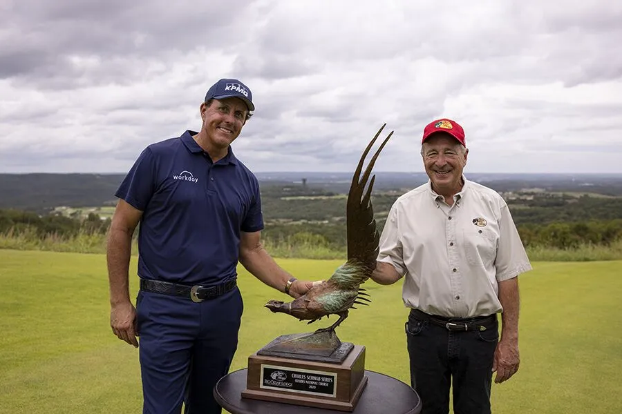 phil mickelson wears dark blue golf attire while standing next to the champsion trophy at ozarks national golf course. He is joined by Johnny Morris wearing dark pants, a white button shirt and red bass pro shops hat