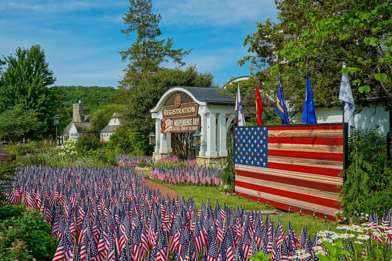 Entrance to a registration area decorated for Independence Day, with hundreds of small American flags, a large wooden flag display, military branch flags, and a banner reading “Happy Independence Day – Land of the Free, Home of the Brave,” surrounded by gardens and trees.