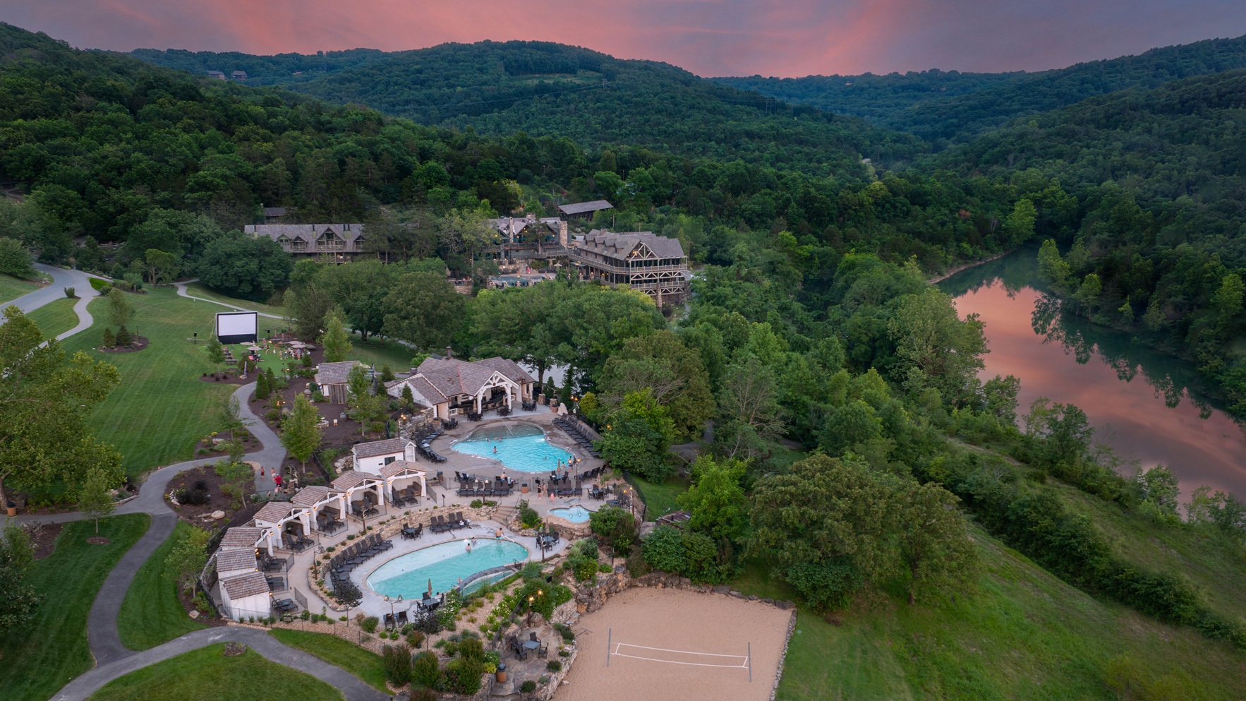 A mountain resort at sunset showcases multiple swimming pools, lodging, and sand volleyball