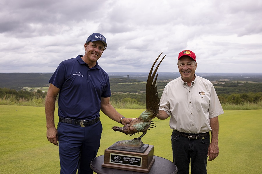 phil mickelson wears dark blue golf attire while standing next to the champsion trophy at ozarks national golf course. He is joined by Johnny Morris wearing dark pants, a white button shirt and red bass pro shops hat