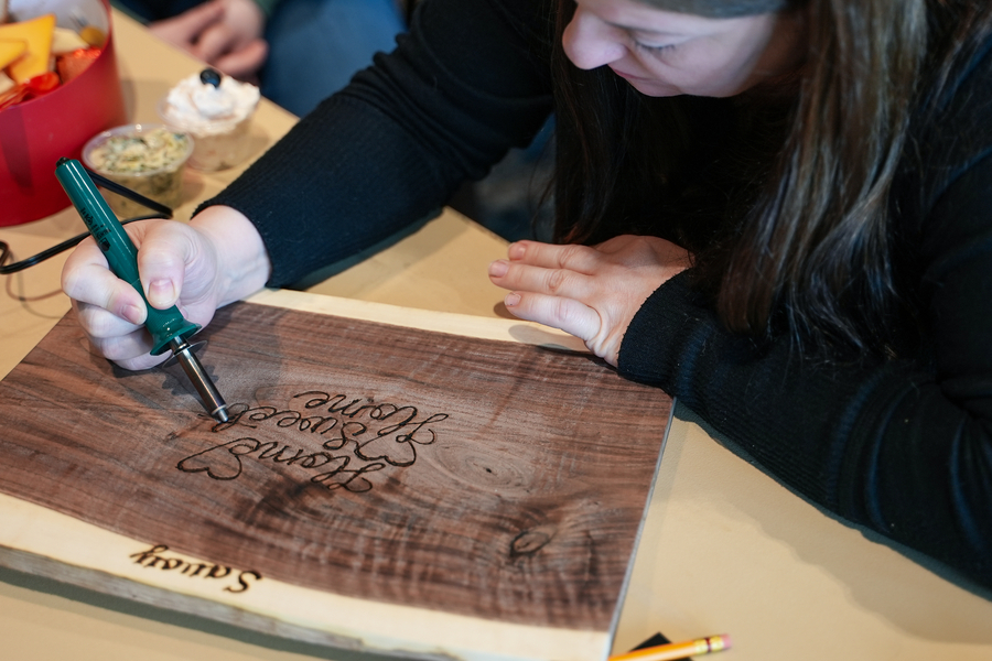 A woman uses a woodburning pen on a charcuterie board to create a whimsical design.