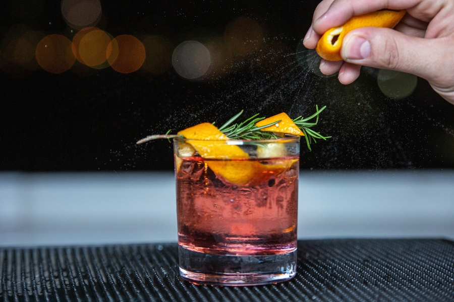A bartender garnishes a cocktail with orange mist and rosemary.