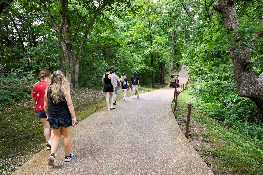 A group of people walk along a paved trail under a green canopy of trees with a single golf cart driving down the trail in the distance.