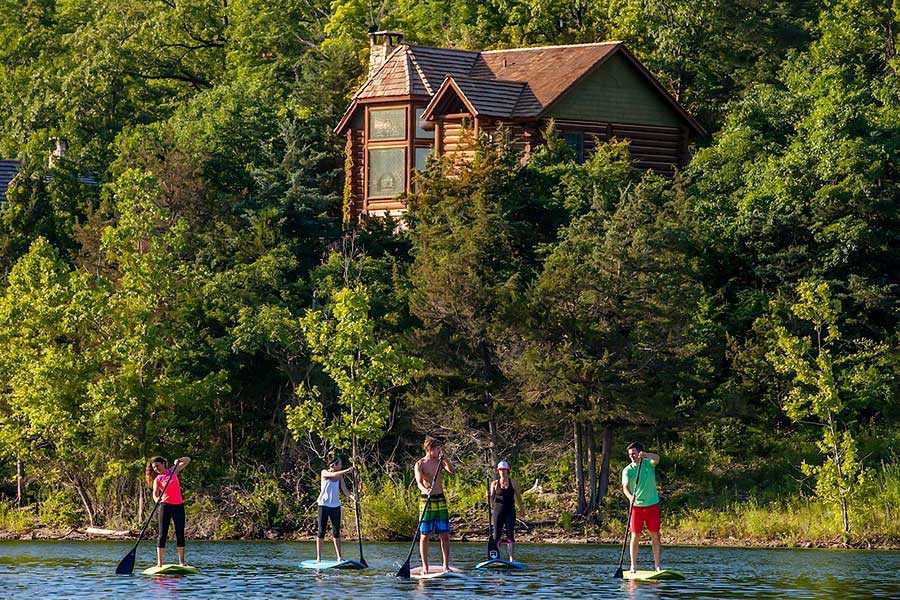 Guests paddleboarding on Table Rock at Big Cedar Lodge, with a Cabin jutting out of the trees behind