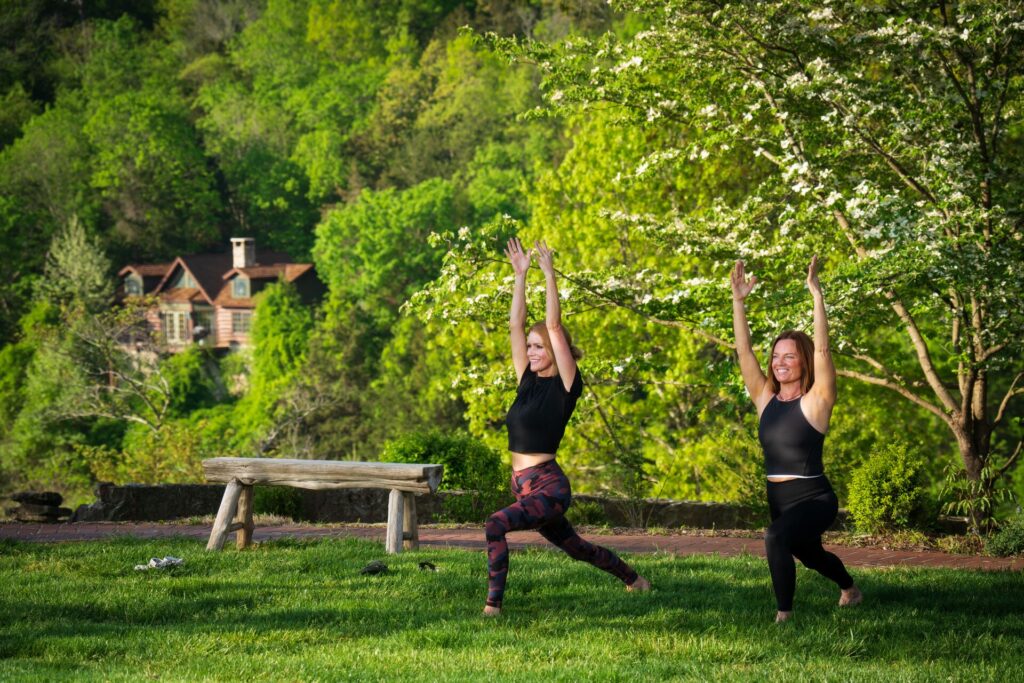 two women practice yoga outside near a walking path surrounded by lush greenery