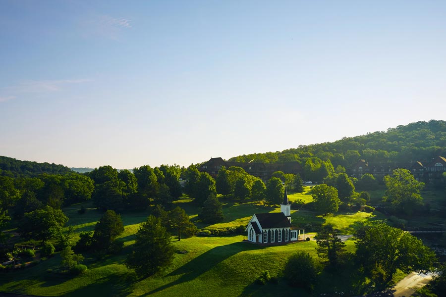 High-angle view of a small, white wooden chapel with a tall steeple, nestled on a lush green hillside surrounded by dense trees at Big Cedar Lodge during a sunny day.