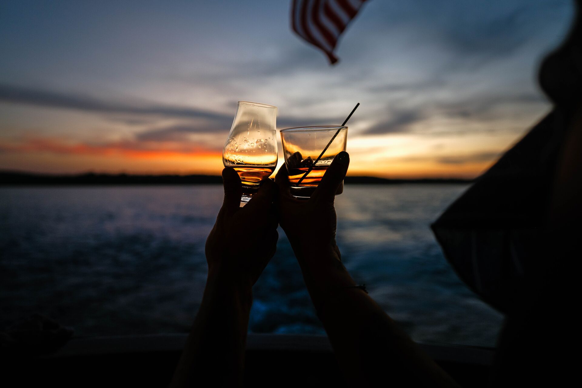 couple toasting with bourbon glasses at dusk overlooking lake from deck of yacht