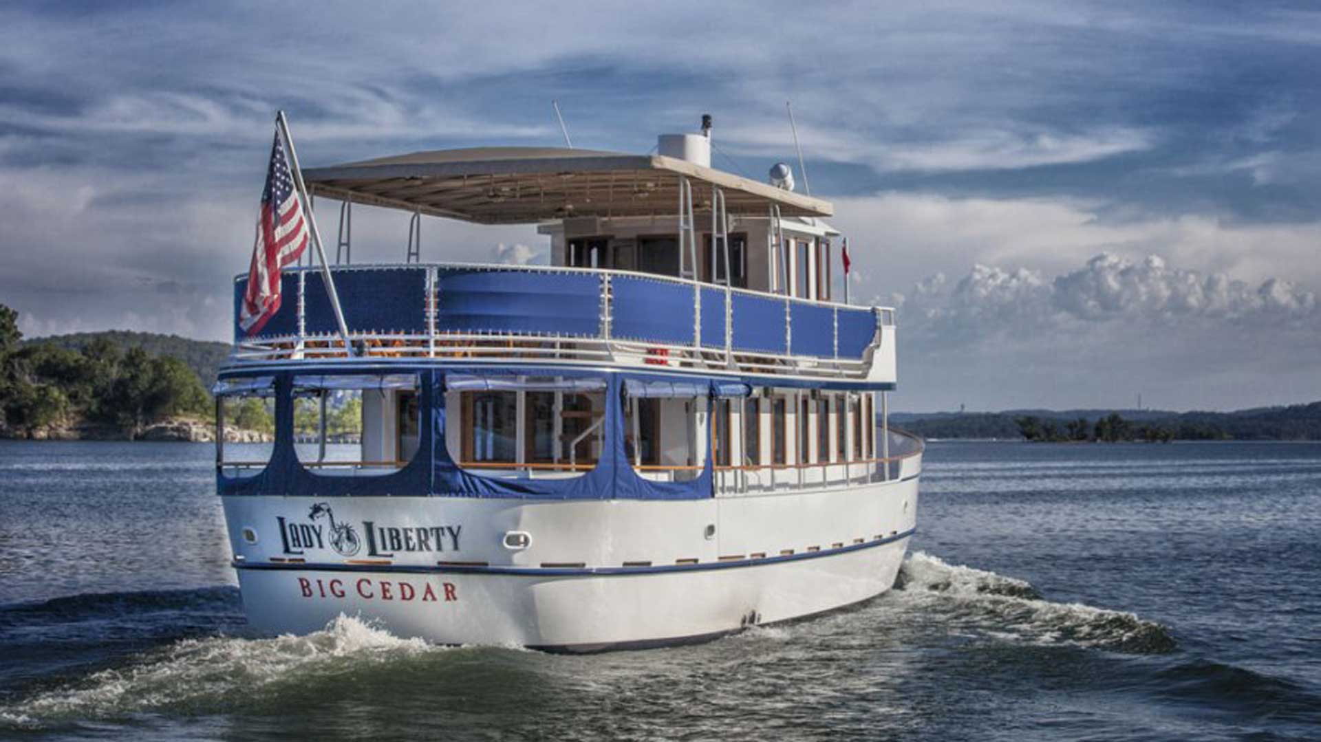 A large white and blue cruise boat named "Lady Liberty" from Big Cedar Lodge sailing across a calm lake. An American flag flies from the stern, and the background shows rolling green hills under a dynamic, cloudy blue sky.