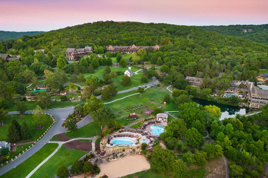High-angle aerial view of a sprawling resort property featuring lodges, swimming pools, lawns, and a small white chapel surrounded by forested hills.