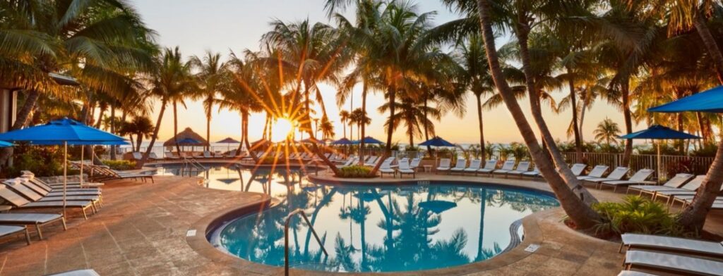 Swimming pool at sunset surrounded by palm trees, basking chairs, and blue umbrellas