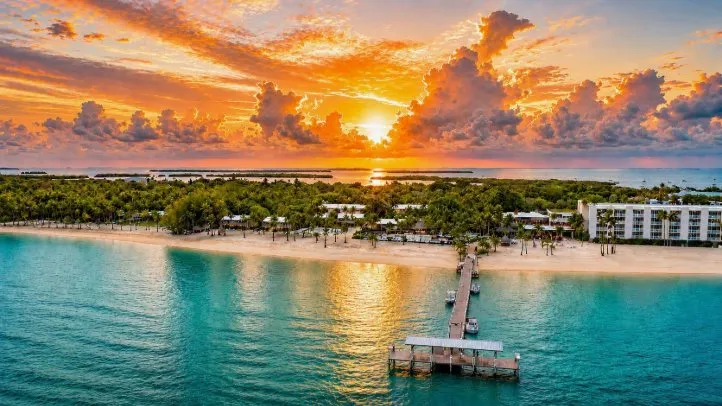 A colorful sunset bursts through large clouds on over a beach resort with many trees and a long wooden pier jutting out from a beach over turquoise water