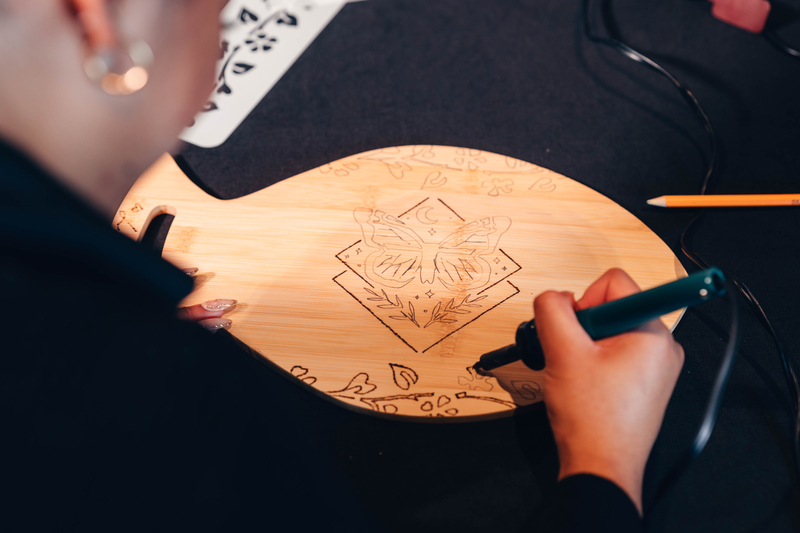 Woman uses a heated pyrography pen to scorch an artistic design of a Moth surrounded by geometric patterns, celestial bodies, and flowering vines