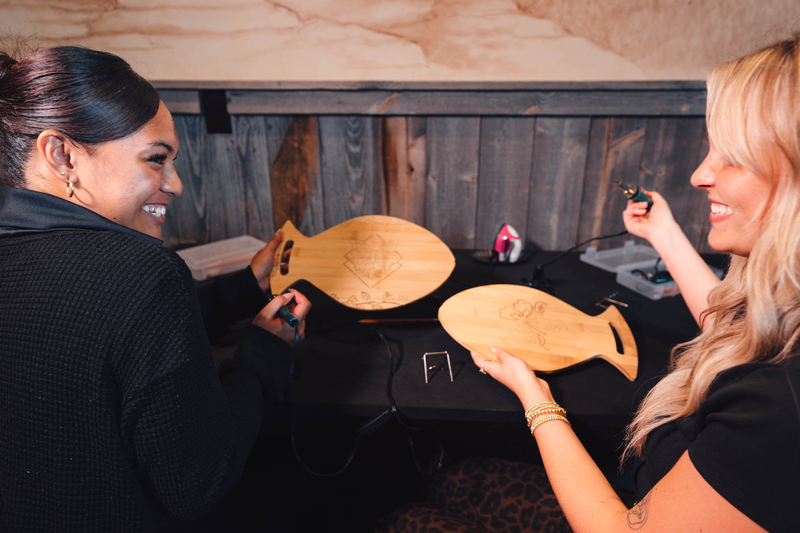 Two friends smile a show each other the wood burned design on a pair of fish shaped cutting boards that they are working on in the activity center at Fun Mountain