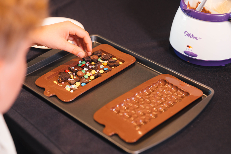 child sprinkles candy and cookie crumbles into a mould for custom chocolate bar crafting next to a pot of tempered melted chocolate