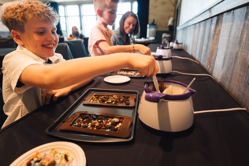 child ladles tempered chocolate from a melting pot into a mould filled with candy and crushed cookies to make a custom chocolate bar crafting