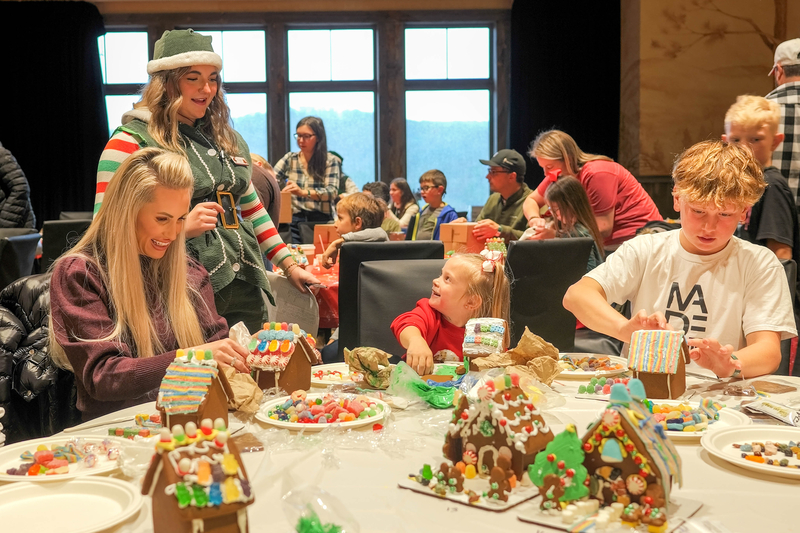 People making any covered gingerbread houses under the supervision of a smiling woman dressed like one of Santa's elves.