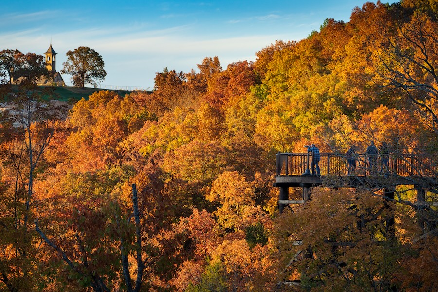 People on a platform overlooking a valley in fall with the stone chapel at Top of the Rock on a hill in the distance