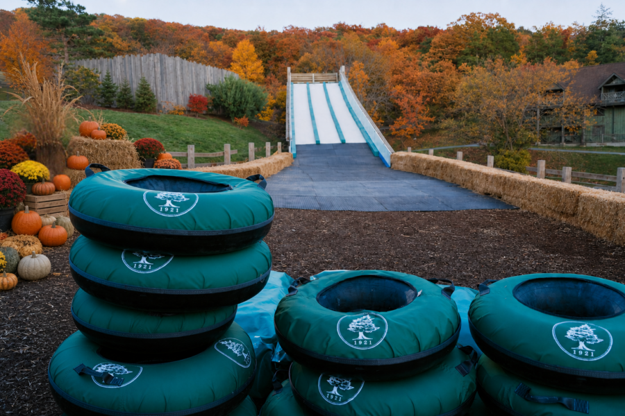 Stacks of dark green inflatable innertubes in front of 4 rows of long white slides enclosed by bales of hay and rounded by autumn trees in the distance