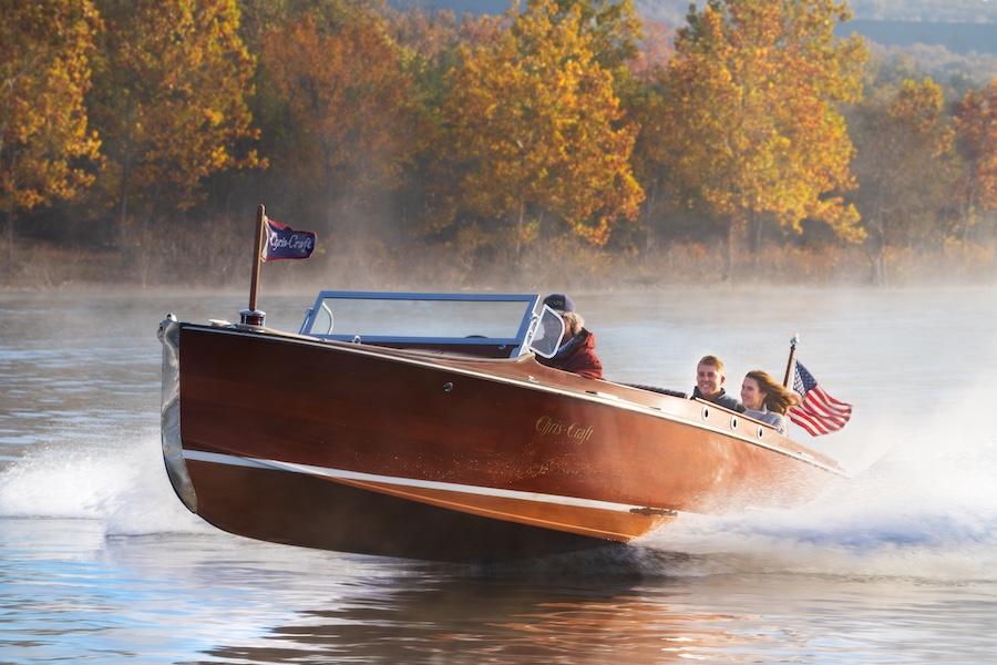 Couple rides vintage wooden speed boat piloted by a captain in the fall with yellow trees in the background