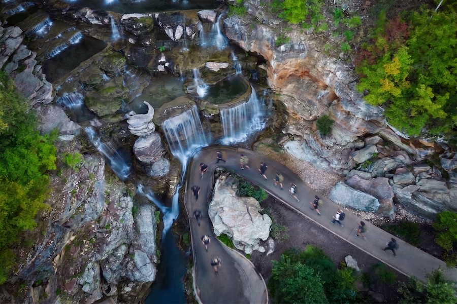 A bird's eye view of people walking with rucking backpacks in a single file line next to a waterfall