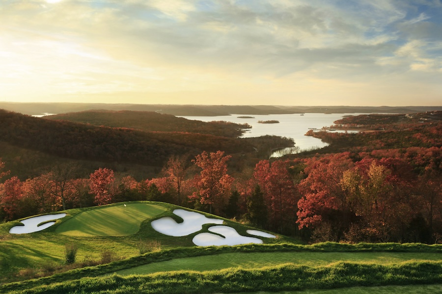 A green golf course overlooking a lake at sunset with hills and valleys cloaked in forests filled with autumn reds and oranges