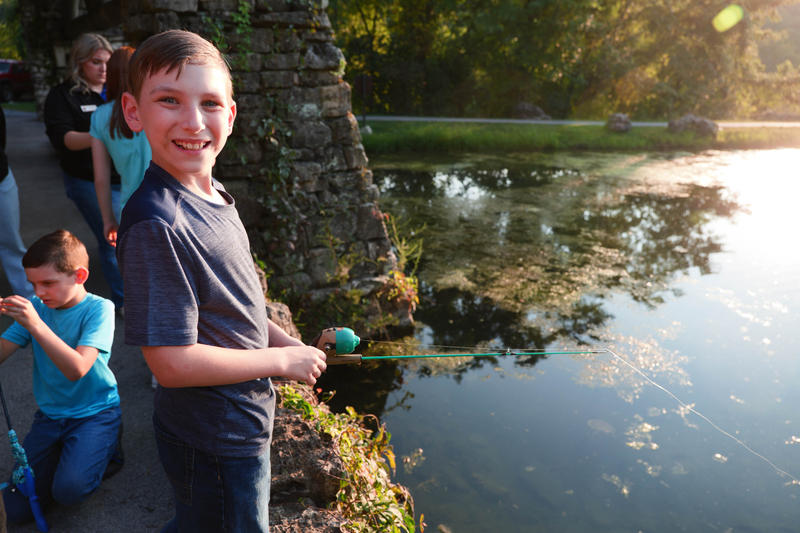 A boy smiles at the camera while reeling in his catch at the fishing pond
