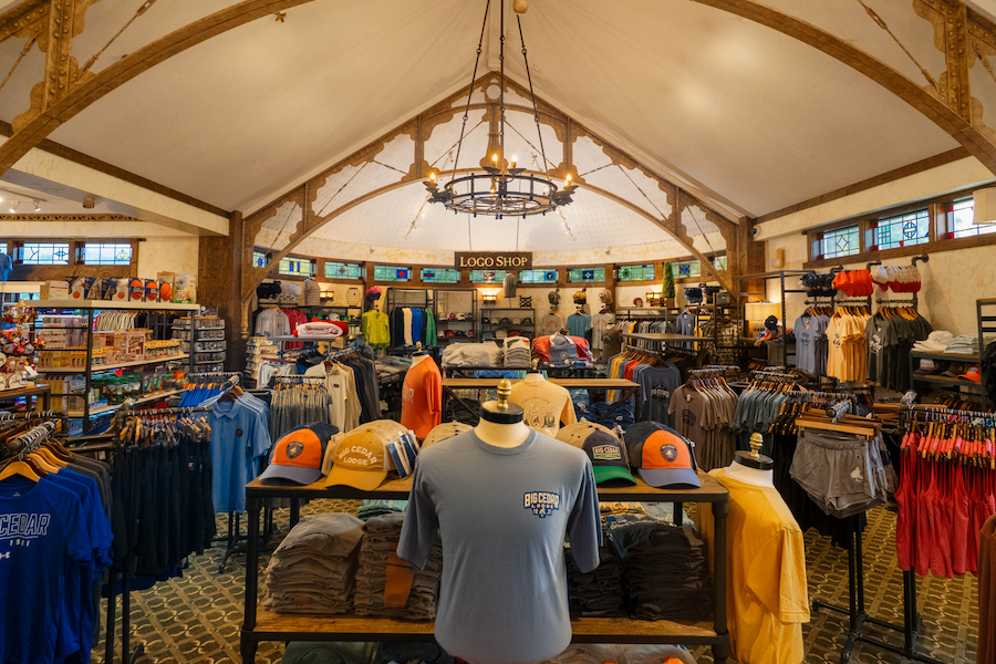 A gift Shop filled with Clothing Displays of T-shirts and Outdoor fashion under a vaulted ceiling with log supports and a rustic chandelier