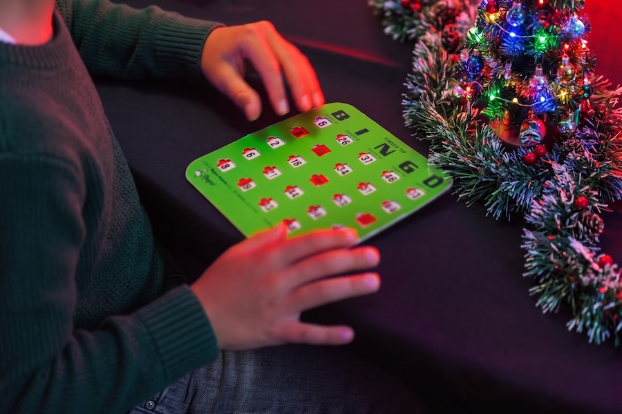 A boy fills out a green bingo card on a table covered with tinsel and Christmas lights.