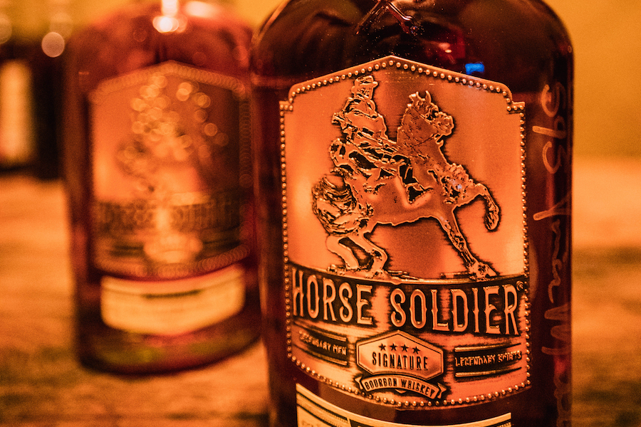 A glass bottle of bourbon with the a metal plate fastened to it with A man riding a rearing horse on top of the words Horse Soldier bourbon