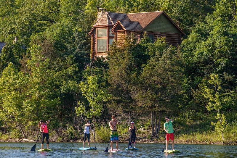 5 people standing on surfboard-like platforms floating on the water with a long paddle that allows them to propel themselves along a wooded coastline under a log cabin on a hill in the background