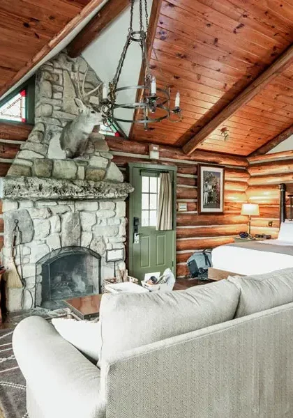 Rustic log cabin interior with a stone fireplace, vaulted wood ceiling, a white sofa, and a large bed.