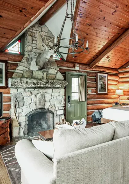 Rustic log cabin interior with a stone fireplace, vaulted wood ceiling, a white sofa, and a large bed.