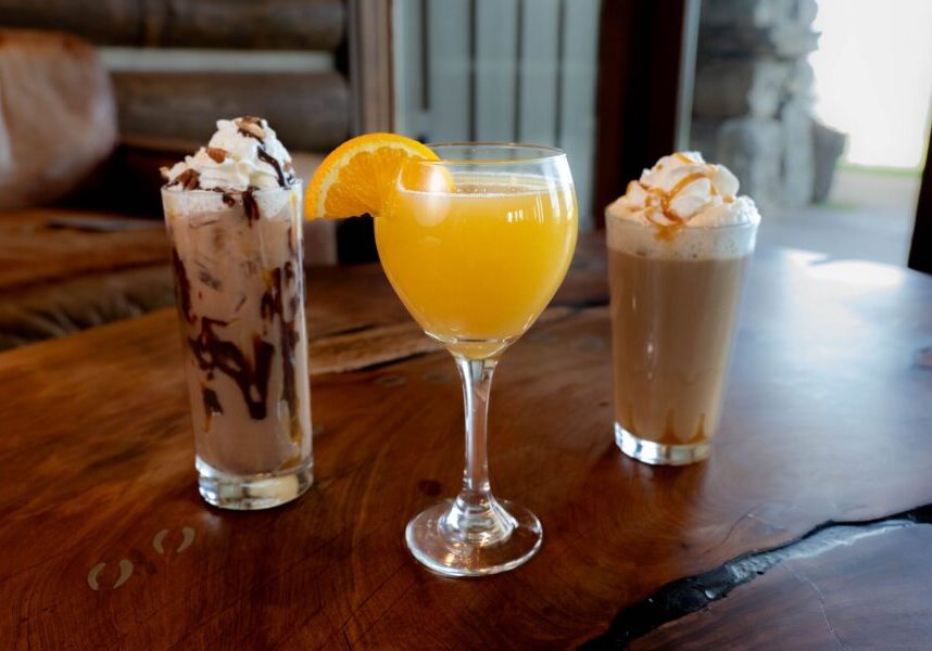 two fancy coffee beverages sit next to a mimosa with orange sticking out of the glass