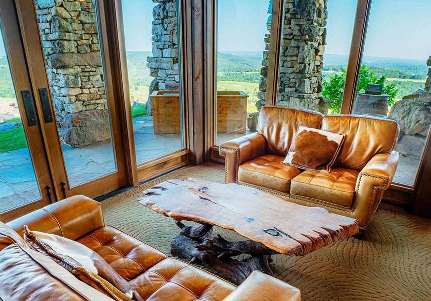 a wooden table, tan leather couches sit next to floor to ceiling windows overlooking the ozark mountains