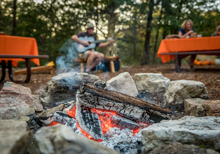 Close up picture of a bonfire with a man playing guitar in the background