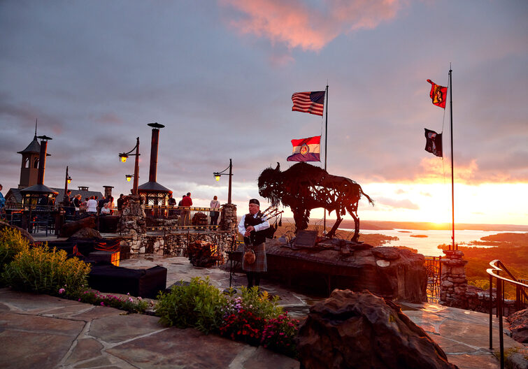 Man Playing Bag Pipes at sunset next to a metal sculpture of a bison
