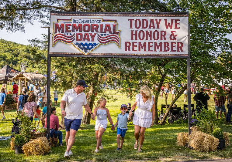 A smiling family walks beneath the Memorial Day sign. People in the background occupy a field with hay blocks and camping chairs. There are several food booths in the distance.