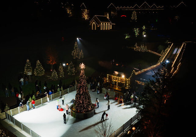 An aerial shot of a square ice rink with a large Christmas tree in the center. People skate and lean agains the railing. A set of four long parallel slides slope down towards a small building outlined with Christmas lights. the entire scene is surrounded by gold, red, and green Christmas lights on trees, and buildings nearby like the Garden Chapel and a big outdoor lodge in the distance.